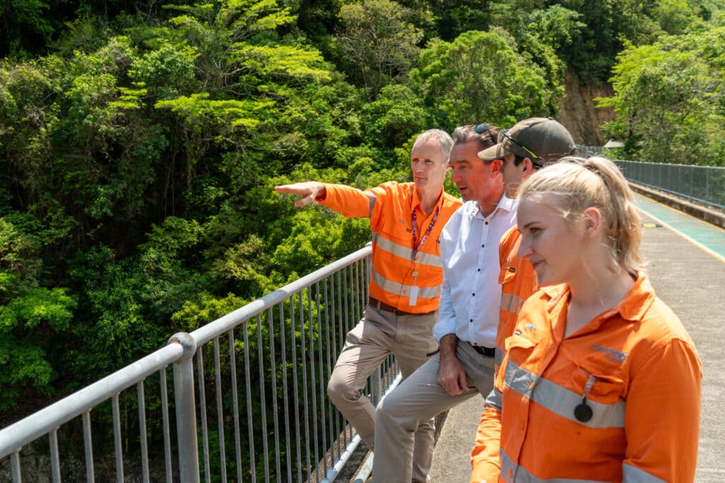 A group photo of CleanCo workers and a local politician. They are standing on the bridge at the Barron Gorge Power Station, pointing and looking out over the Barron River, wearing their safety gear.