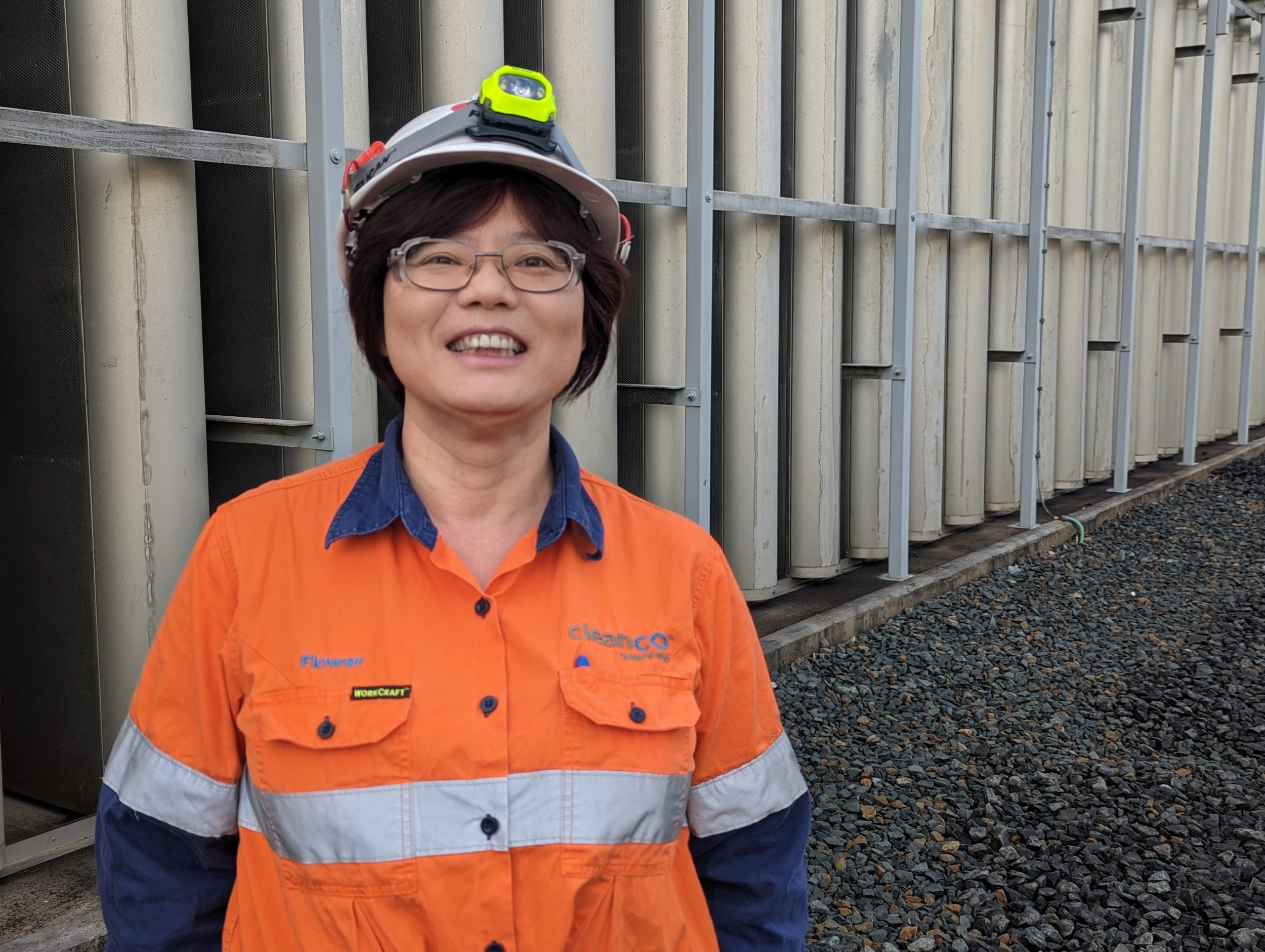 A photo of a smiling CleanCo employee named Flower, wearing orange high visibility clothing and a hard hat, standing in front of a gas fired power station.