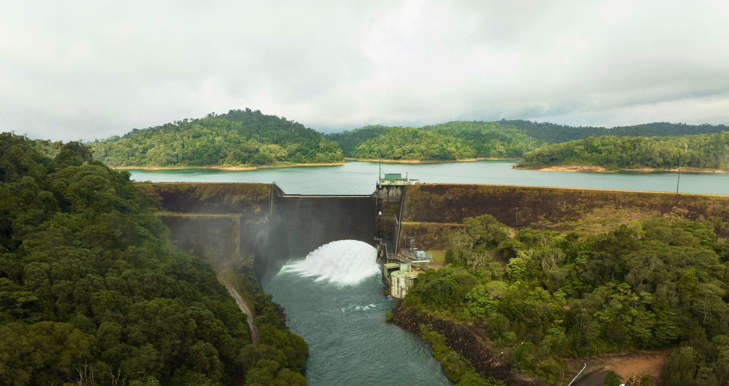 An aerial view of the Koombooloomba dam wall. It is surrounded by green hills. The dam wall is opened slightly allowing water to gush through.