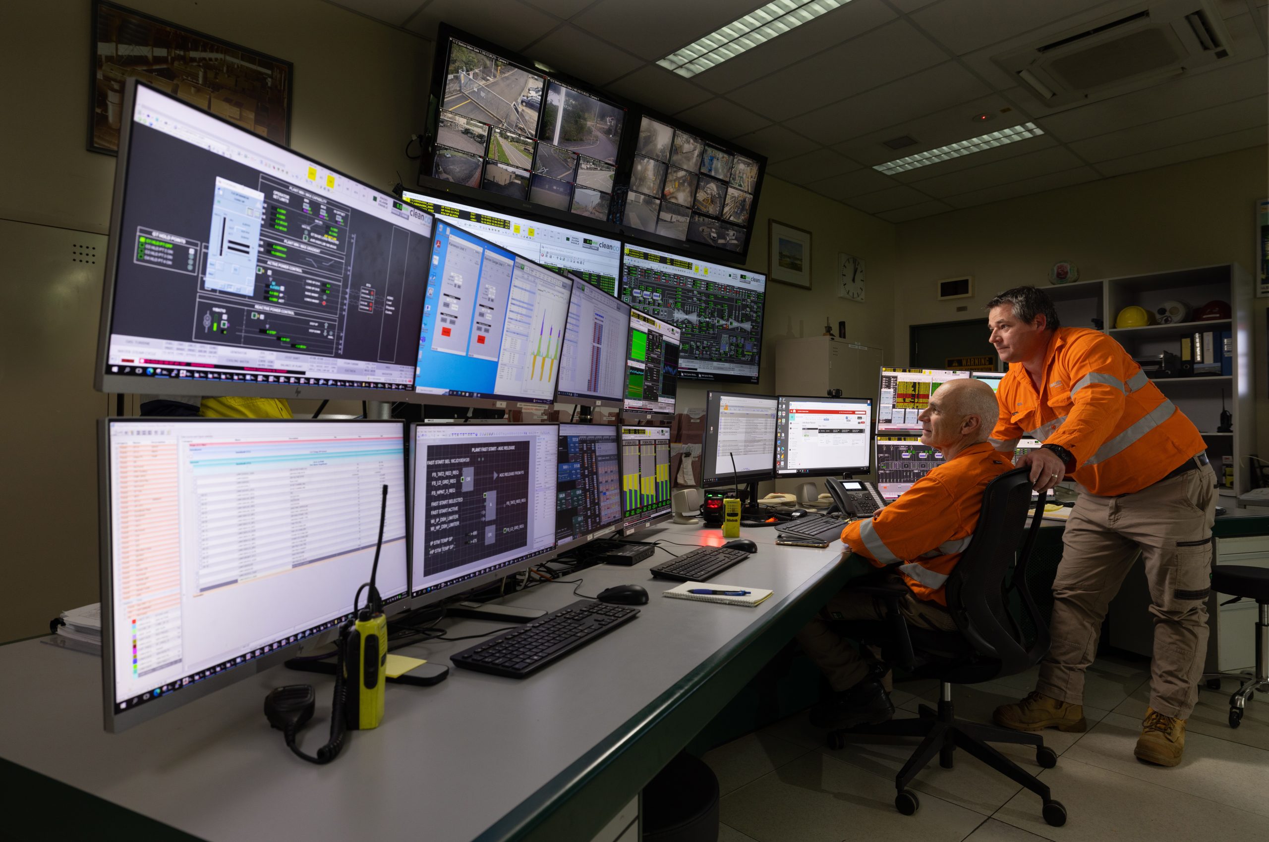 A photograph of two CleanCo workers at the Swanbank Power Station. They are wearing safety gear and staring at the many screens in the control room.