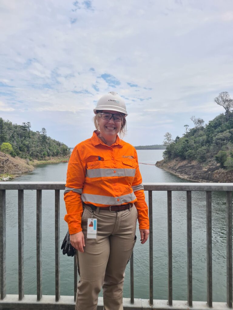 A photograph of a CleanCo worker wearing high visibility uniform, smiling, with Wivenhoe Dam behind her.